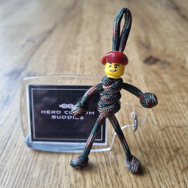May include: A handmade keychain featuring a Lego figure with a red beret and a camouflage-patterned body. The keychain includes a clear tag with the text "HERO CUSTOM BUDDIES". The figure is approximately 7.6 cm tall.