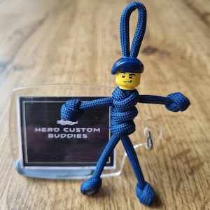 May include: A navy blue keychain featuring a Lego figure with a yellow head and a blue beret. The figure is attached to a clear plastic tag with the text "HERO CUSTOM BUDDIES" in white. The figure's body is made of woven blue cord.