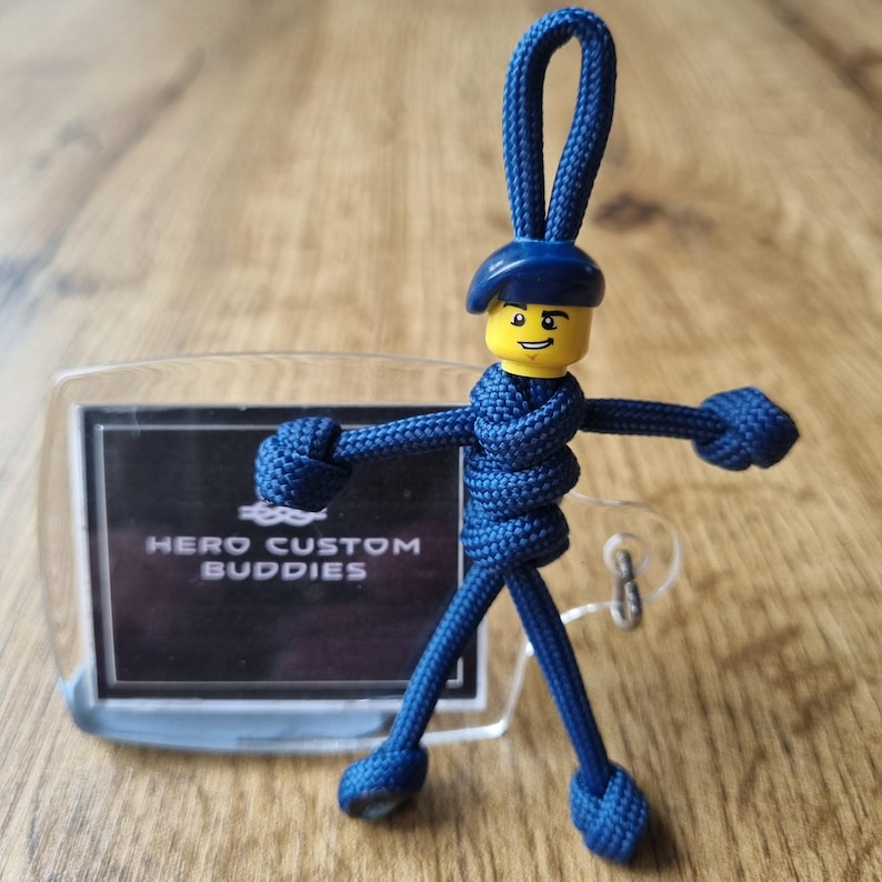 May include: A navy blue keychain featuring a Lego figure with a yellow head and a blue beret. The figure is attached to a clear plastic tag with the text "HERO CUSTOM BUDDIES" in white. The figure's body is made of woven blue cord.