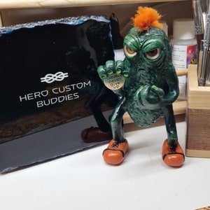 May include: Green pickle-shaped figurine with orange hair, holding a heart-shaped charm that says "Never never give up". The figurine is standing on a white surface.