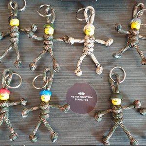 May include: A selection of handmade keyrings. Each keyring features a small figure with a yellow head and a different coloured hat. The figures are made from camouflage-patterned cord and have metal key rings. The Hero Custom Buddies logo is visible.