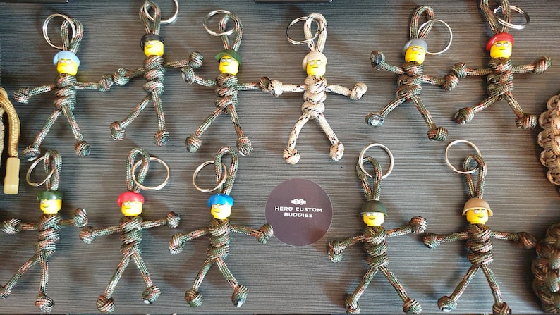 May include: A selection of handmade keyrings. Each keyring features a small figure with a yellow head and a different coloured hat. The figures are made from camouflage-patterned cord and have metal key rings. The Hero Custom Buddies logo is visible.