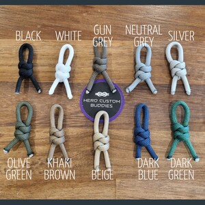 May include: A variety of colors of paracord, tied in a figure eight knot, with the text "HERO CUSTOM BUDDIES" on a white circle. The colors include black, white, gun grey, neutral grey, silver, olive green, khaki brown, beige, dark blue, and dark green.