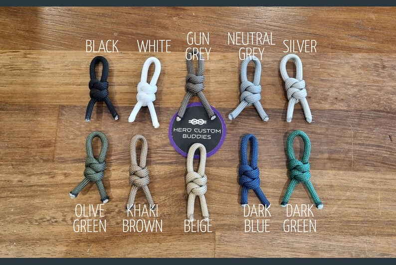 May include: A variety of colors of paracord, tied in a figure eight knot, with the text "HERO CUSTOM BUDDIES" on a white circle. The colors include black, white, gun grey, neutral grey, silver, olive green, khaki brown, beige, dark blue, and dark green.