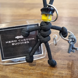 以下が含まれることがあります： 黒いパラコードのキーホルダーフィギュアで、黄色のレゴの頭が黒い帽子をかぶっています。フィギュアは銀色の鍵のセットを持っています。キーホルダーは、透明なプラスチック製のカードホルダーに取り付けられており、そこに「HERO CUSTOM BUDDIES」という文字が印刷されています。