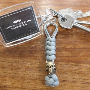 May include: A gray paracord keychain with a skull-shaped bead and a clear plastic keychain tag that says "Hero Custom Buddies".