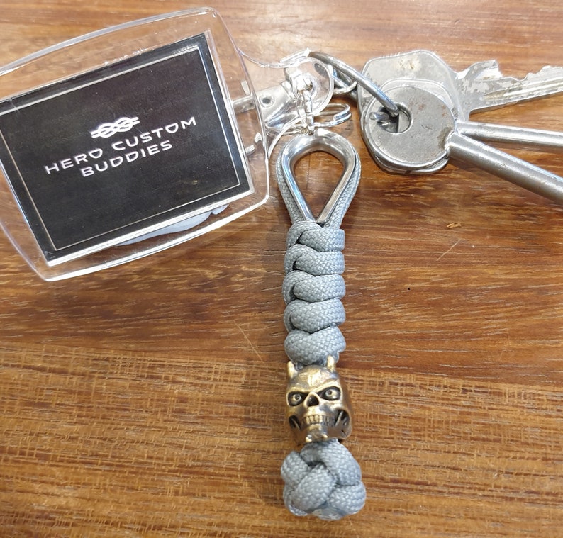 May include: A gray paracord keychain with a skull-shaped bead and a clear plastic keychain tag that says "Hero Custom Buddies".