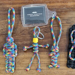 Puede incluir: Cinco llaveros de paracord coloridos, que incluyen un llavero de figura, un llavero de nudo y un llavero de bucle. Los llaveros están hechos con paracord colorido y tienen anillos de llaves plateados. Los llaveros se muestran sobre una superficie de madera con una tarjeta de plástico transparente que dice "Hero Custom Buddies".