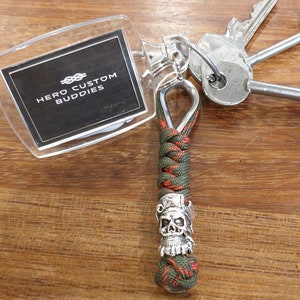 May include: Green and red paracord keychain with a silver skull bead and a clear plastic tag that says "HERO CUSTOM BUDDIES".