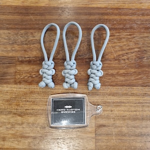 Puede incluir: Tres tiradores de cremallera de paracord gris con un diseño anudado. Los tiradores de cremallera están dispuestos en una fila sobre una superficie de madera. Una etiqueta de plástico transparente con el texto "HERO CUSTOM BUDDIES" está debajo de los tiradores de cremallera.