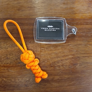 May include: Orange paracord keychain with a knot in the shape of a small figure. The keychain is attached to a clear plastic tag with the text "HERO CUSTOM BUDDIES" printed in white.