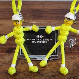 以下が含まれることがあります： 白いハードハットと黄色のレゴの顔をした、黄色のパラコードのフィギュア2体。フィギュアは手をつないでおり、透明なプラスチックカードに「HERO CUSTOM BUDDIES」という文字が書かれています。