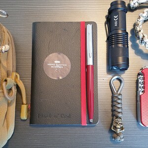 May include: A brown canvas bag with a zipper and a small yellow figure keychain. Next to it is a black notebook with the text "Hero Custom Journals" and a red pen. A black flashlight, a red pocketknife, and a gray paracord keychain with a skull charm are also visible.