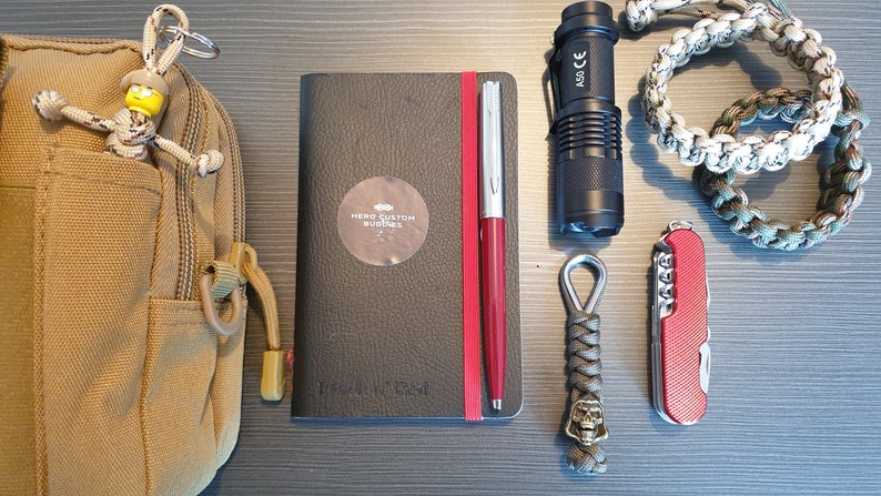 May include: A brown canvas bag with a zipper and a small yellow figure keychain. Next to it is a black notebook with the text "Hero Custom Journals" and a red pen. A black flashlight, a red pocketknife, and a gray paracord keychain with a skull charm are also visible.