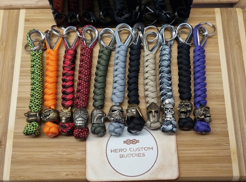 May include: A collection of 12 colorful paracord keychains with different metal bead charms. The charms include a skull, a helmet, a Star Wars character, and a dragon. The keychains are displayed on a wooden surface with a business card that reads "Hero Custom Buddies".