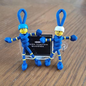 May include: Two keychains made of blue paracord with yellow accents, each featuring a yellow plastic head with a printed face and a helmet. One figure wears a blue helmet, the other a white helmet. A black sign reads HERO CUSTOM BUDDIES.