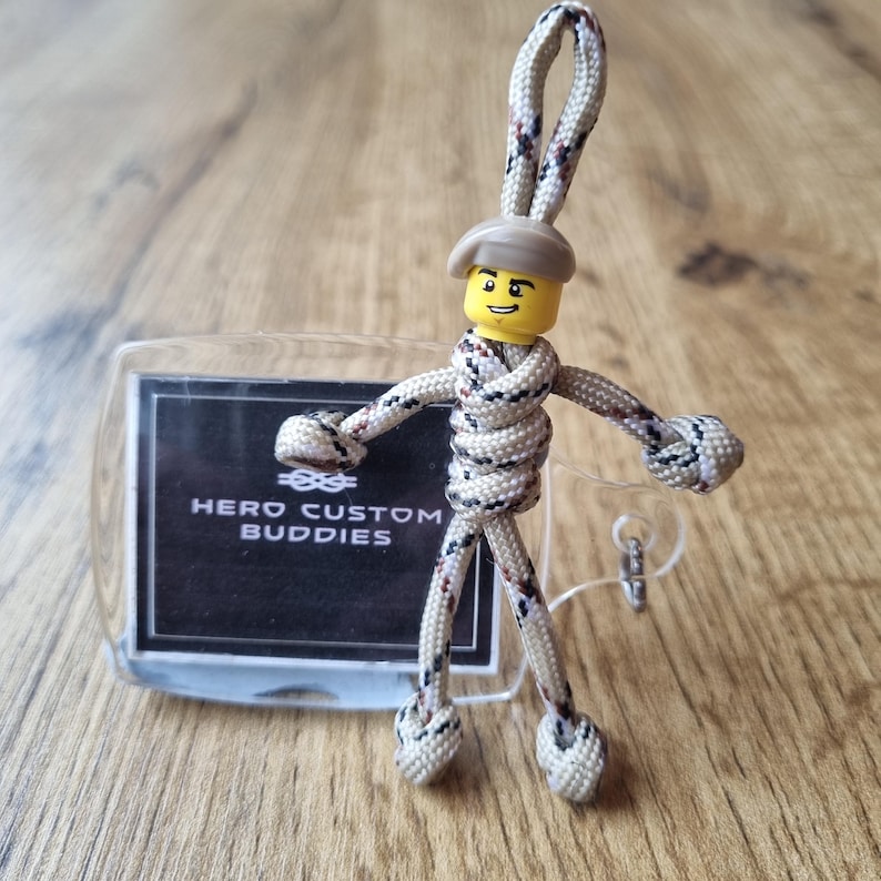May include: A handmade keychain featuring a Lego figure head with a beige hat, integrated into a woven rope design. The rope is beige with black and red accents. A clear tag with the text "HERO CUSTOM BUDDIES" is also included.