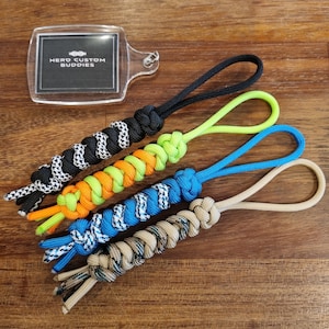 May include: Four colourful paracord keychains in black and white, neon green and orange, blue and white, and tan and brown. A clear acrylic keychain with the text "HERO CUSTOM BUDDIES" is also present. The keychains are on a wooden surface.