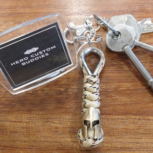 May include: A tan and brown paracord keychain with a silver metal loop and a silver metal Spartan helmet charm. The keychain is attached to a clear acrylic keychain tag with the text "HERO CUSTOM BUDDIES" in black.