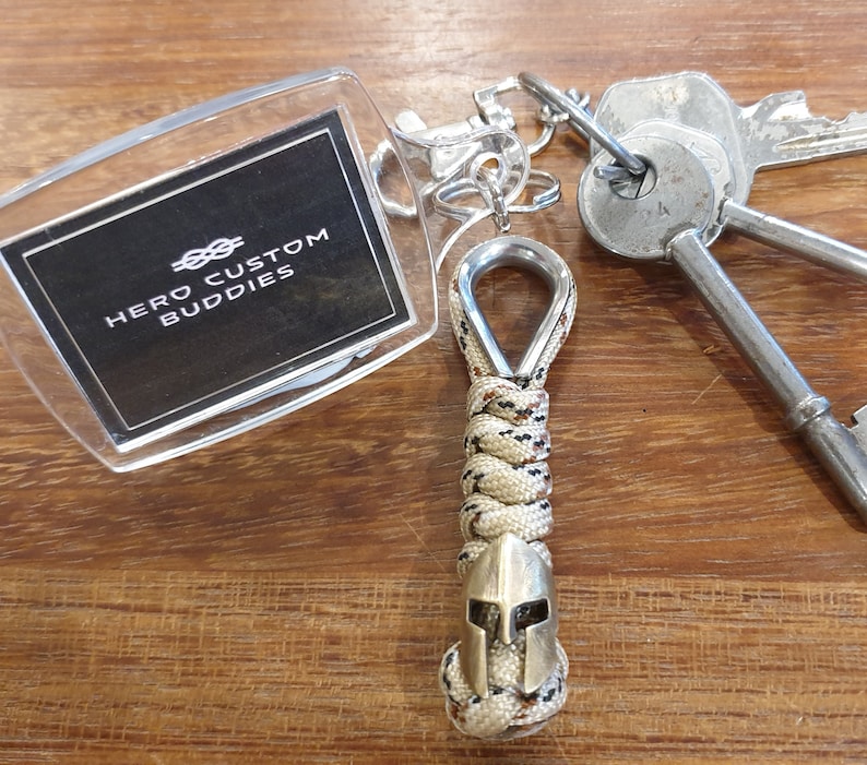 May include: A tan and brown paracord keychain with a silver metal loop and a silver metal Spartan helmet charm. The keychain is attached to a clear acrylic keychain tag with the text "HERO CUSTOM BUDDIES" in black.