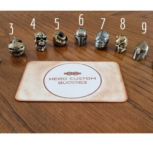 May include: Eleven metal keychains featuring various characters, including a skull, pumpkin, Mandalorian, Spider-Man, and Darth Vader. The keychains are arranged in a row on a wooden surface with the text "HERO CUSTOM BUDDIES" on a card in the center.