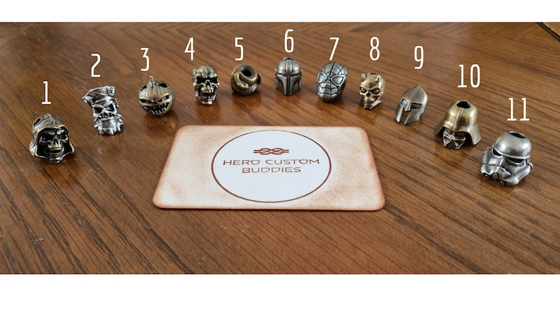 May include: Eleven metal keychains featuring various characters, including a skull, pumpkin, Mandalorian, Spider-Man, and Darth Vader. The keychains are arranged in a row on a wooden surface with the text "HERO CUSTOM BUDDIES" on a card in the center.
