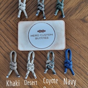 May include: A selection of paracord accessories in shades like beige, army camo, olive green, khaki brown, desert camo, coyote brown camo, and navy blue. A card with the text "HERO CUSTOM BUDDIES" is also visible.