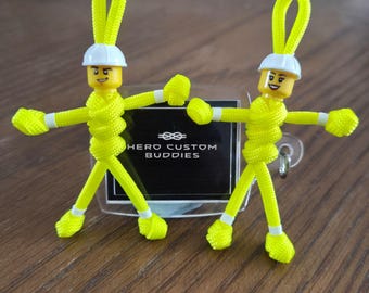 May include: Two neon yellow paracord figures with white hard hats and yellow plastic heads featuring printed facial expressions. Each figure has a loop at the top and stands in front of a black rectangular sign with the text HERO CUSTOM BUDDIES.