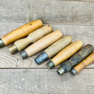 May include: A collection of six vintage tool handles made of various shades of light to dark brown wood, each featuring a cylindrical shape and a metal ferrule at the base. The handles vary in length, thickness, and wood grain texture.