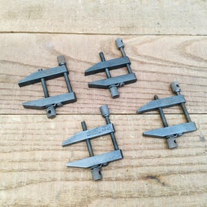 May include: Four dark grey metal Eclipse No. 410 toolmaker clamps arranged in a group. Each clamp features two parallel jaws connected by two threaded rods with knurled adjustment knobs at the ends for tightening and securing small workpieces.