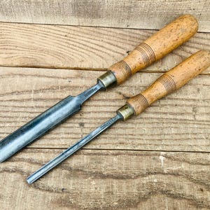 Peut inclure: Deux gouges à bois avec des lames en acier poli et des manches en bois brun clair. Chaque outil comporte une virole en laiton reliant la lame au manche, qui présente des anneaux décoratifs gravés près du sommet. Les lames sont courbées.