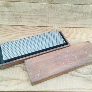 May include: A rectangular sharpening stone with a gray, textured surface, set in a wooden tray. The wooden lid is next to the tray. The stone is used for sharpening knives and other tools. The wood has a natural, aged appearance.