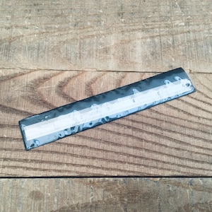 May include: A black ruler with white markings and numbers, lying on a wooden surface. The ruler appears to be approximately 6 inches long, with clear, easy-to-read measurements.