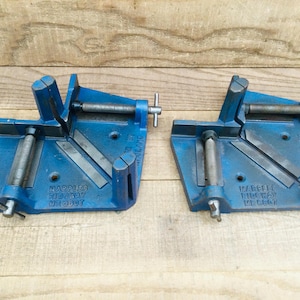 May include: Two blue metal corner clamps for woodworking, each featuring two threaded steel rods with tightening handles. The base of each clamp is embossed with the text Marples Ridgway M2607 and Made in England, designed for securing joints.