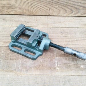 May include: A teal-colored metal drill press vise featuring a rectangular base with elongated mounting slots, a stationary jaw, a movable jaw, and a long threaded screw handle with a cylindrical end piece for manual adjustment and tightening.