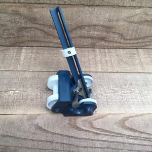 May include: A blue and white mechanical tool featuring a long, slotted arm extending upward from a base equipped with four white wheels. The device includes a metallic adjustment lever and a white sliding collar positioned along the central blue arm.