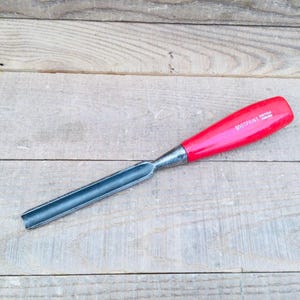 May include: A woodworking chisel with a red handle and a silver blade. The handle has the text "FOOTPRINT SHEFFIELD ENGLAND" printed on it. The chisel is lying on a light-coloured wooden surface.