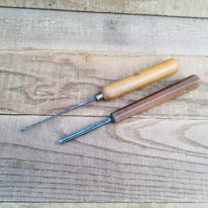 May include: Two woodworking tools with metal shafts and wooden handles. One tool features a long, thin, cylindrical metal rod with a light-colored wooden handle, while the other has a shorter, grooved metal blade with a dark-colored wooden handle.