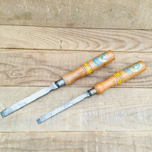 May include: Two wood chisels with light brown wooden handles and silver metal blades. Each handle features a circular blue and gold label with text and three thin yellow rings near the metal ferrule. The blades are flat and rectangular.