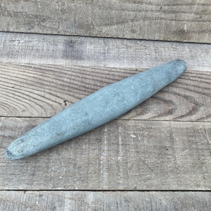 May include: A long, slender, cylindrical sharpening stone with a matte, blue-grey finish. The stone features a tapered, rounded shape that is thicker in the centre and narrows towards both ends, displaying a natural, weathered, and textured surface.