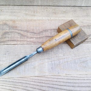 May include: A wood carving chisel with a wooden handle and a metal blade. The chisel is resting on a small wooden block, with the blade angled towards the viewer. The handle is light brown, and the blade is silver.