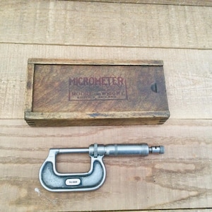 May include: A vintage micrometer and its wooden storage box. The box is brown with the word "MICROMETER" and the brand name "MOORE & WRIGHT" printed on it. The micrometer is silver and has a curved frame.
