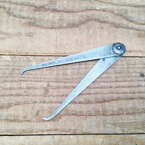 May include: A vintage metal caliper tool with curved tips, open and resting on a wooden surface. The tool is silver-toned and has a rivet at the top, connecting the two arms. The wood grain is visible in the background.