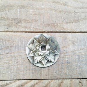 May include: A silver-coloured, circular decorative element with a floral design. The piece features a central square opening surrounded by stylized leaves and floral details. The object is set against a wooden background.