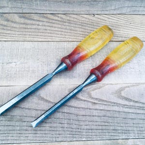 May include: Two wood chisels featuring metallic blades and translucent amber-colored plastic handles that transition into a darker reddish-brown hue near the metal ferrule. The tools have a flat, beveled cutting edge and a rounded grip design.