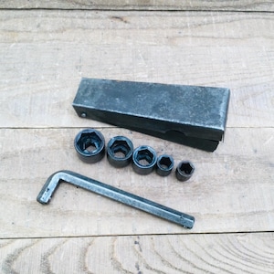 May include: A collection of metal sockets and a wrench displayed on a wooden surface. The sockets are of different sizes, and there is a rectangular metal box. The tools have a used, industrial look.