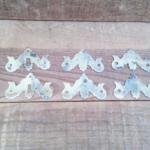 May include: Six antique brass keyhole escutcheons with a decorative, symmetrical design. The escutcheons are tarnished, showing signs of age, and are arranged on a wooden surface with visible grain.