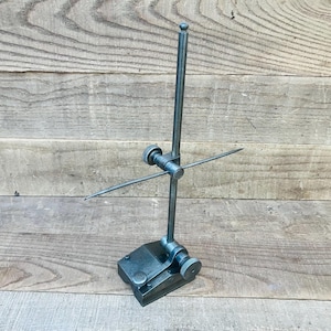 May include: A dark grey metal dial indicator stand featuring a heavy rectangular base, a vertical support rod, and an adjustable horizontal arm with a circular tightening knob. The tool includes a secondary adjustment knob at the base.