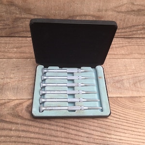May include: A set of six small screwdrivers with silver handles and pointed tips, neatly arranged in a black case with a light blue interior. The case is open, revealing the tools against a wooden background.