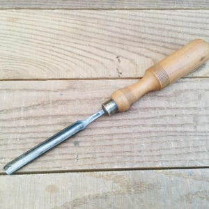 May include: A woodworking chisel with a light brown wooden handle and a silver metal blade. The handle has a cylindrical shape with a slightly wider grip area. The blade is straight and tapers to a cutting edge.